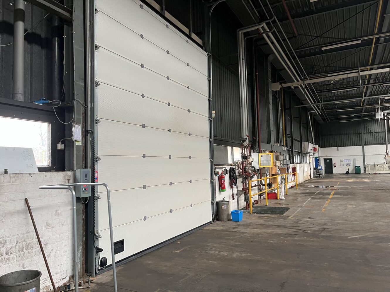 Een industrieel magazijninterieur met een grote gesloten overheaddeur, hoge plafonds, zichtbare leidingen en diverse gereedschappen en apparatuur langs de muren.