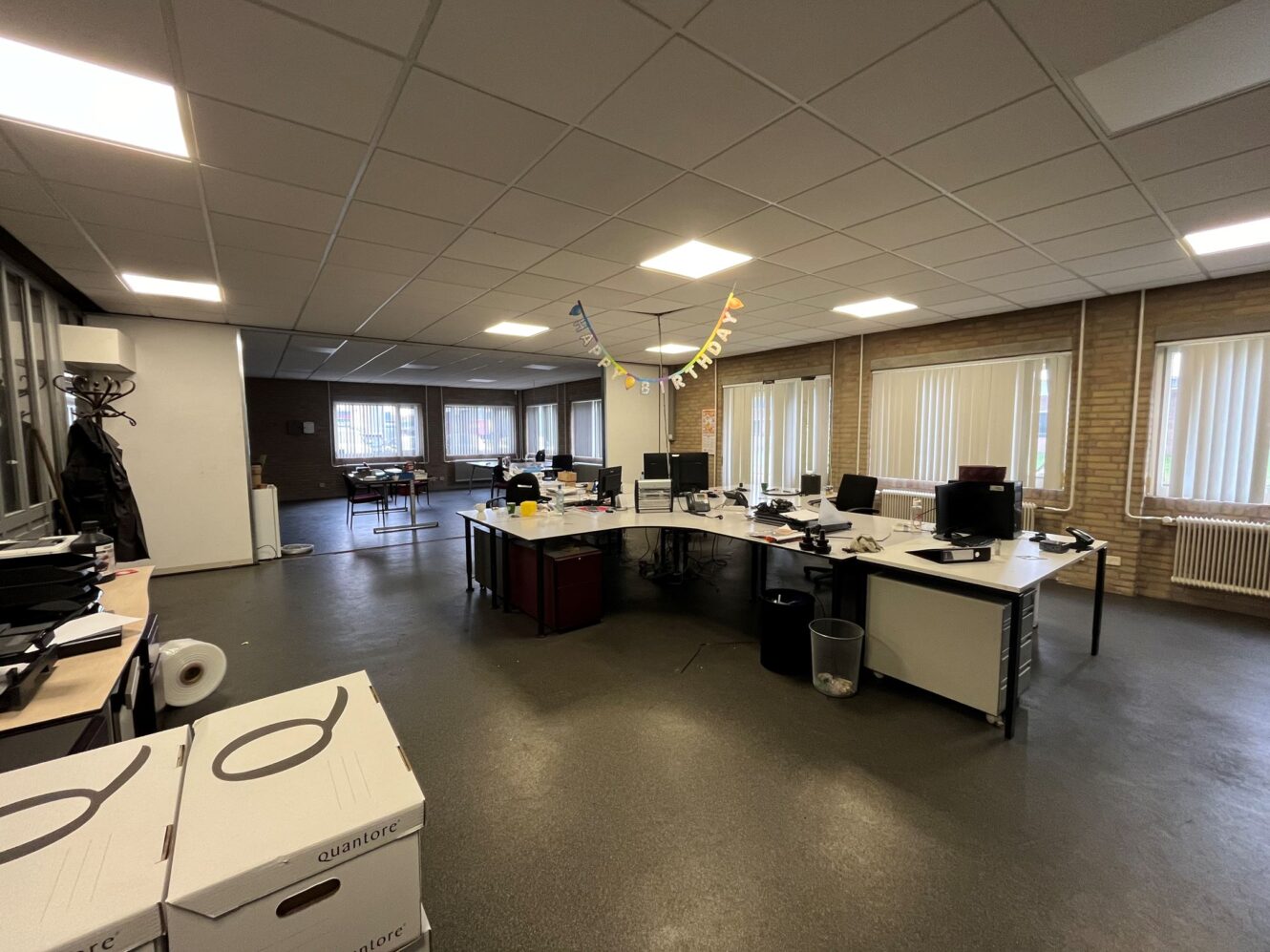 Ruim kantoor met verschillende bureaus, computers, kantoorbenodigdheden en dozen. Een banier met vlaggen hangt boven de bureaus; ramen langs de muren laten natuurlijk licht binnen.