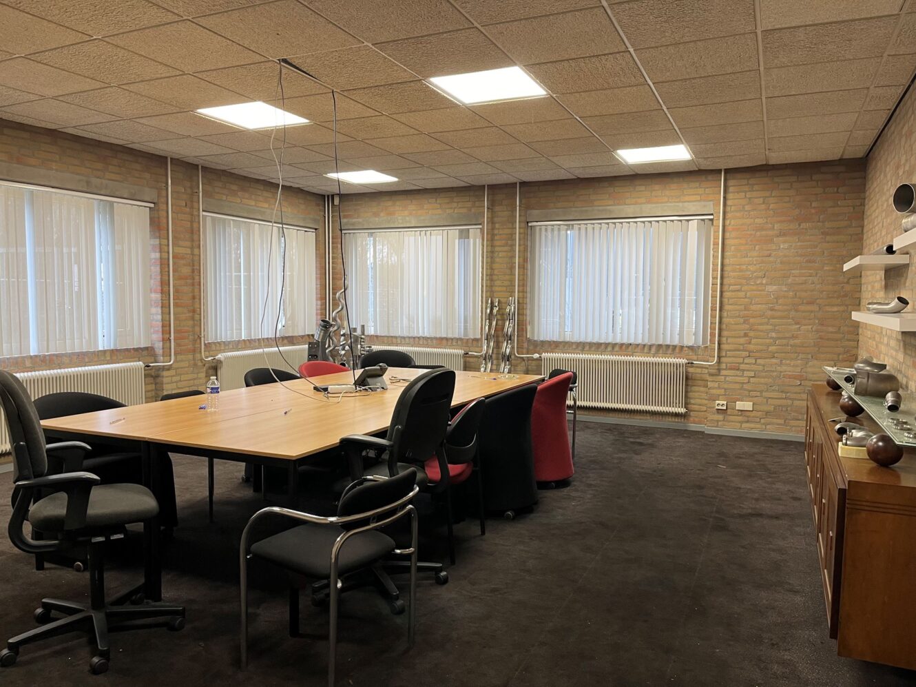 Een vergaderruimte met een grote houten tafel omringd door stoelen, bakstenen muren, ramen met jaloezieën en een dressoir met decoratieve voorwerpen.