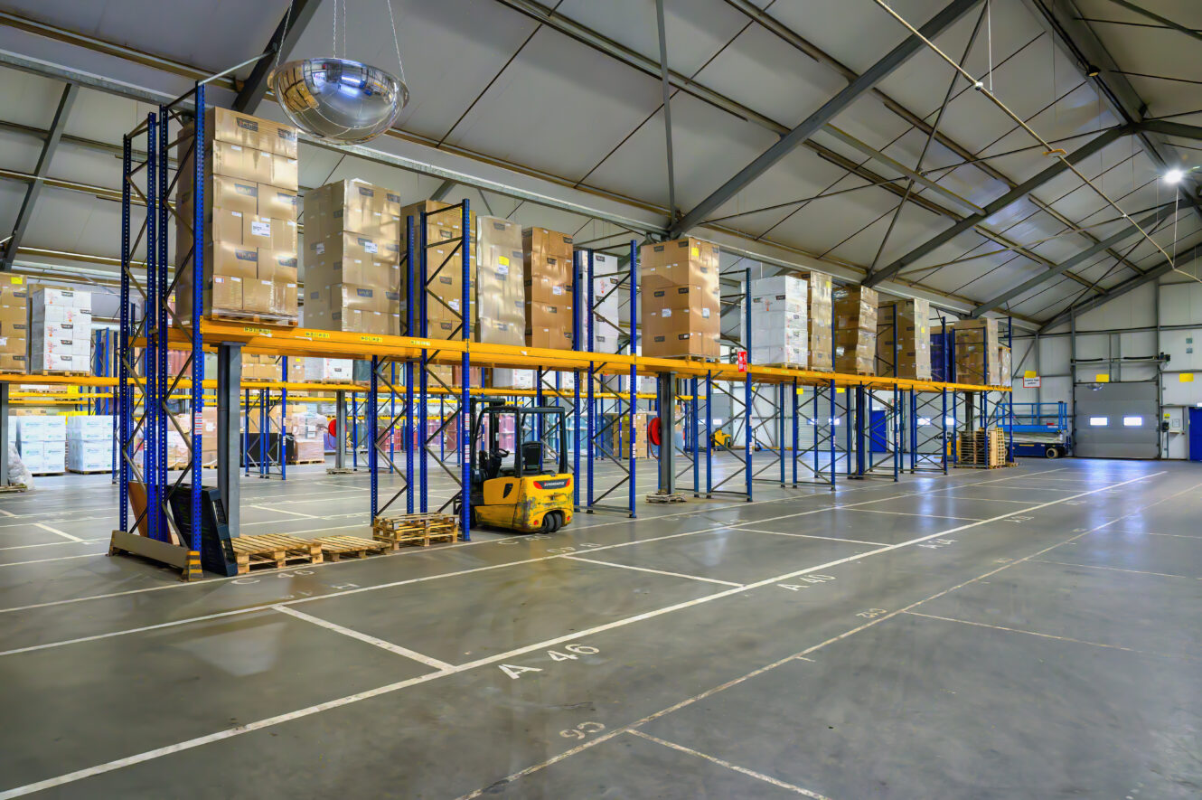 Groot magazijninterieur met hoge rekken vol dozen, een gele vorkheftruck, lege pallets en een discobal die aan het plafond hangt.