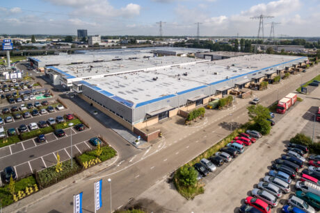 Een groot industrieel magazijncomplex met een blauw en wit dak, omringd door parkeerplaatsen vol auto's en verschillende bestelwagens op aangrenzende wegen.