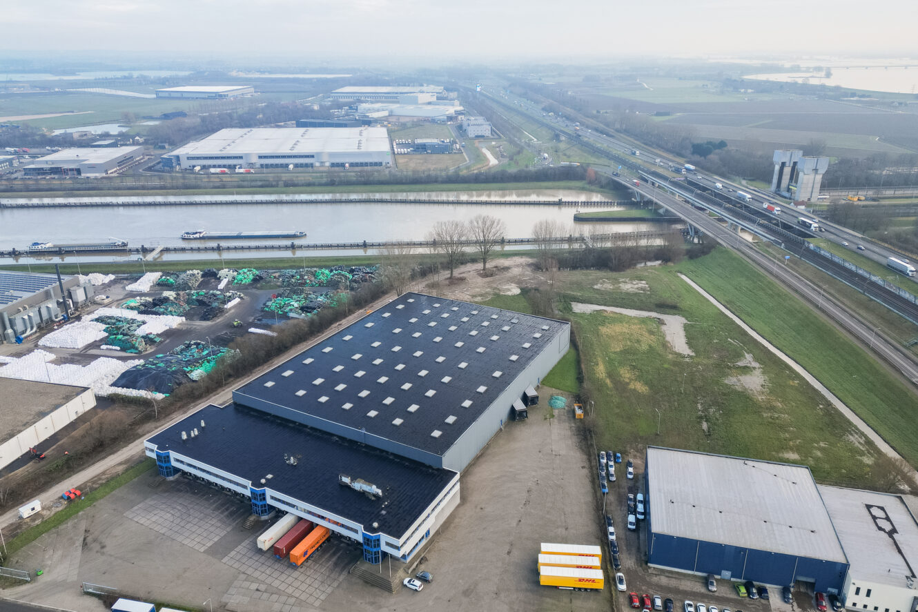 Luchtfoto van een industrieel pakhuiscomplex bij een kanaal, met aangrenzende wegen, geparkeerde voertuigen, groene zones en andere industriële gebouwen op de achtergrond.