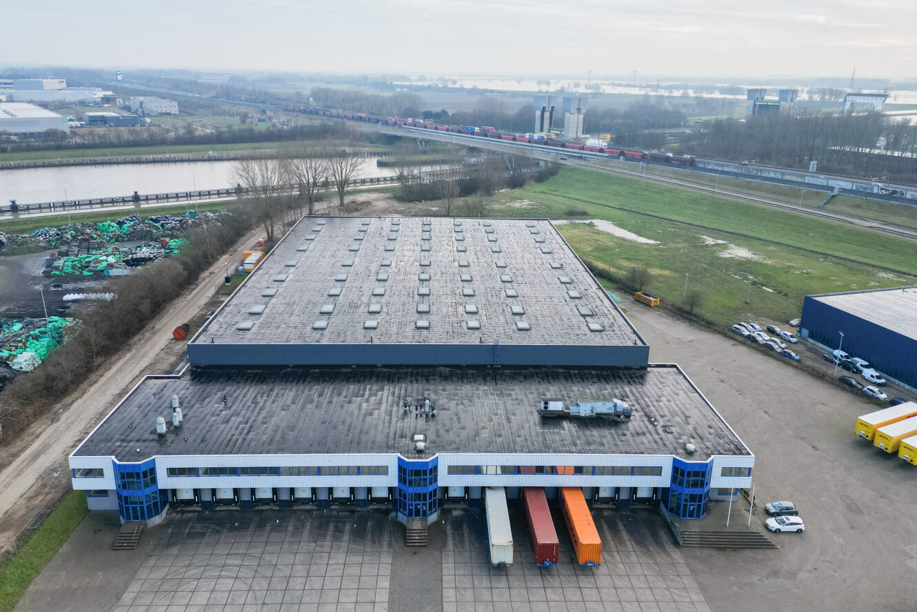 Luchtfoto van een groot industrieel magazijn met meerdere laadkades en verschillende vrachtwagens buiten geparkeerd, omringd door wegen, gras en nabijgelegen gebouwen.