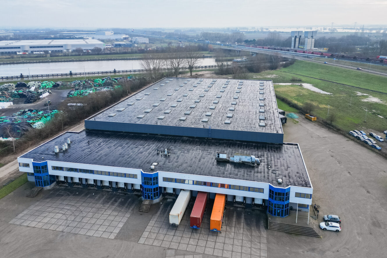 Luchtfoto van een groot magazijn met een plat dak, meerdere laadkades en drie geparkeerde opleggers aan de voorkant, omgeven door geplaveide en met gras begroeide gebieden.