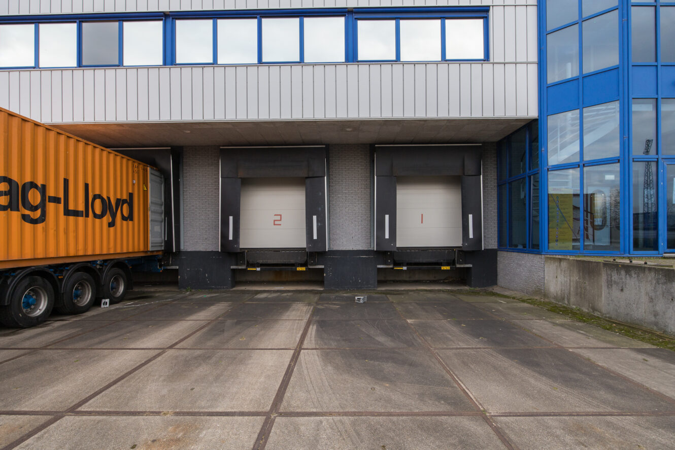 Twee laadkades genummerd 1 en 2 zijn zichtbaar tussen een gebouw en een gele vrachtwagentrailer, met betonnen bestrating op de voorgrond.