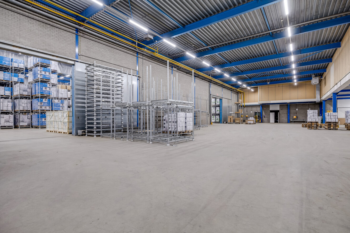 Ruim industrieel magazijninterieur met hoge plafonds, metalen rekken, gestapelde pallets en goederen in dozen langs de muren onder heldere bovenverlichting.