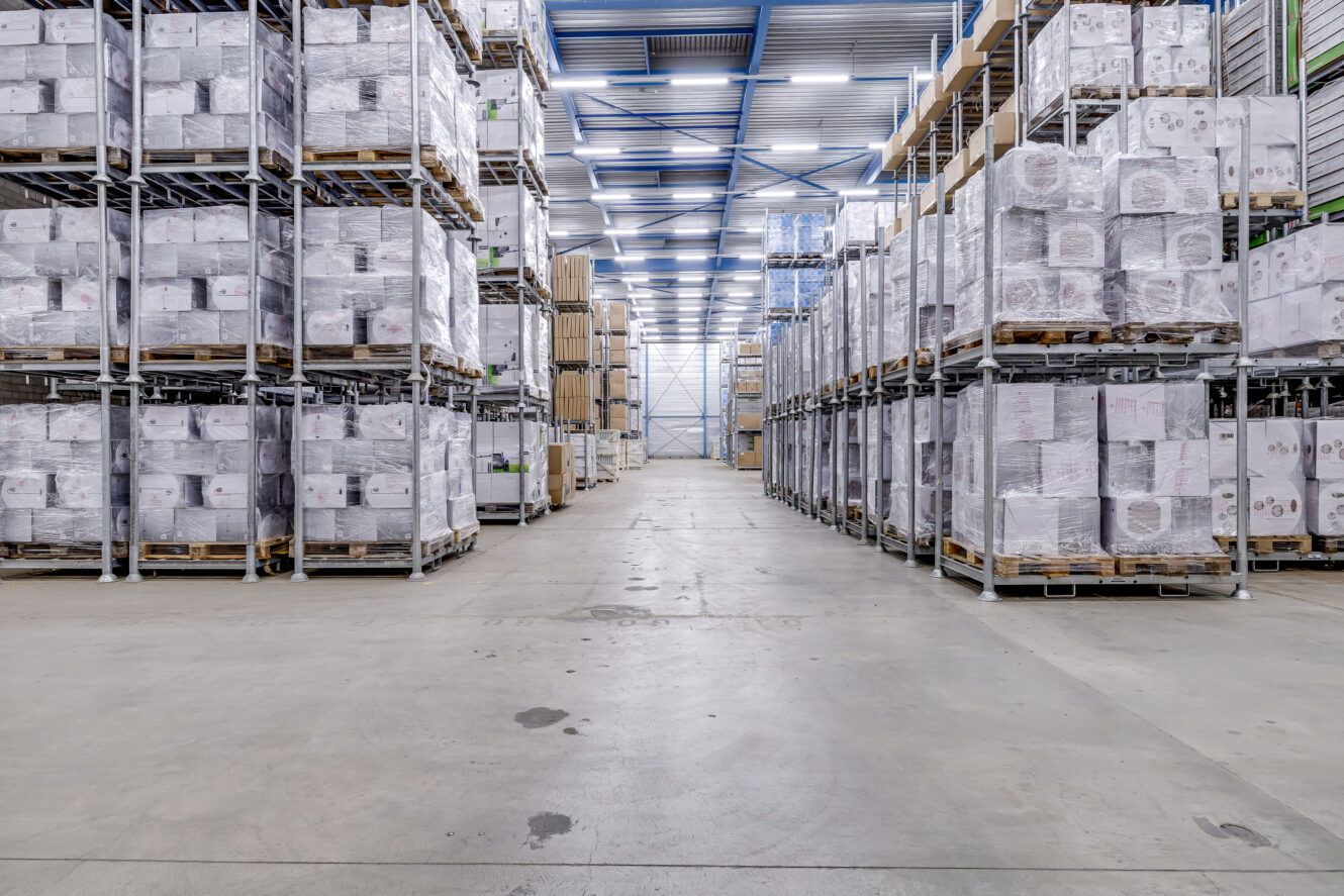 Groot magazijninterieur met rijen hoge metalen rekken volgestapeld met dozen en pallets, onder heldere bovenverlichting en een breed middenpad.