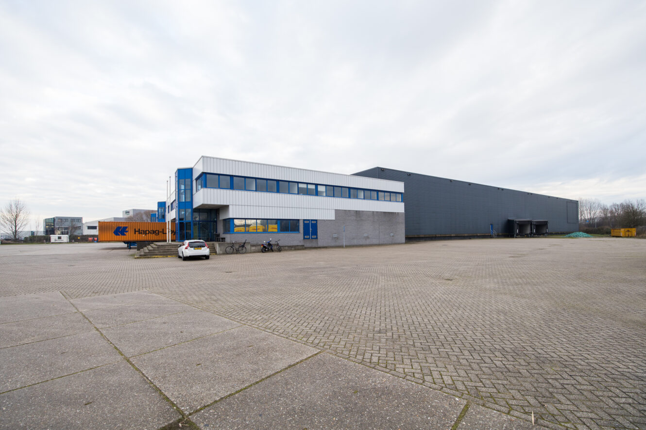 Een groot, modern industrieel gebouw met blauwe accenten en een kleine witte auto buiten geparkeerd op een meestal leeg geplaveid terrein.
