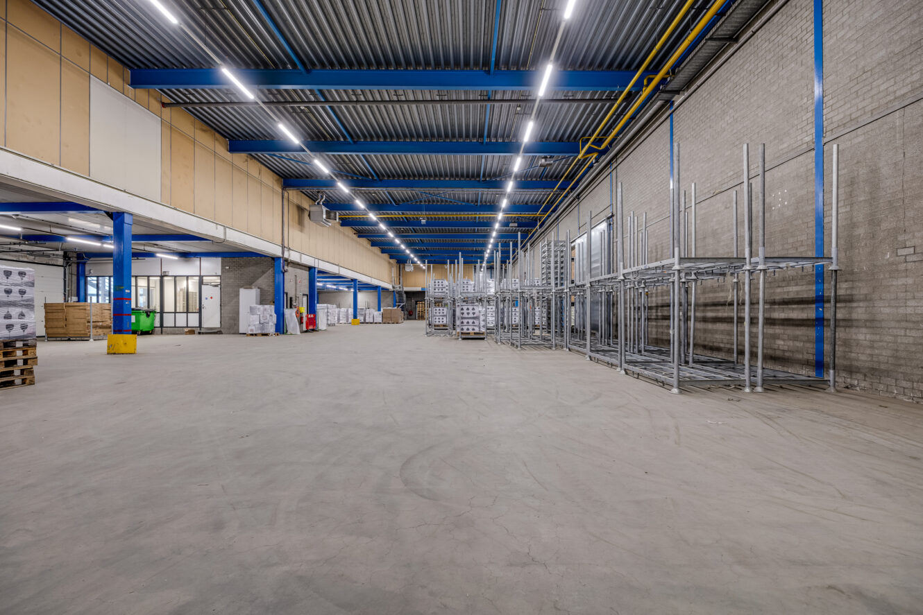 Groot, leeg magazijninterieur met metalen rekken, gestapelde dozen en betonnen vloeren onder blauwe stalen plafondbalken en tl-verlichting.