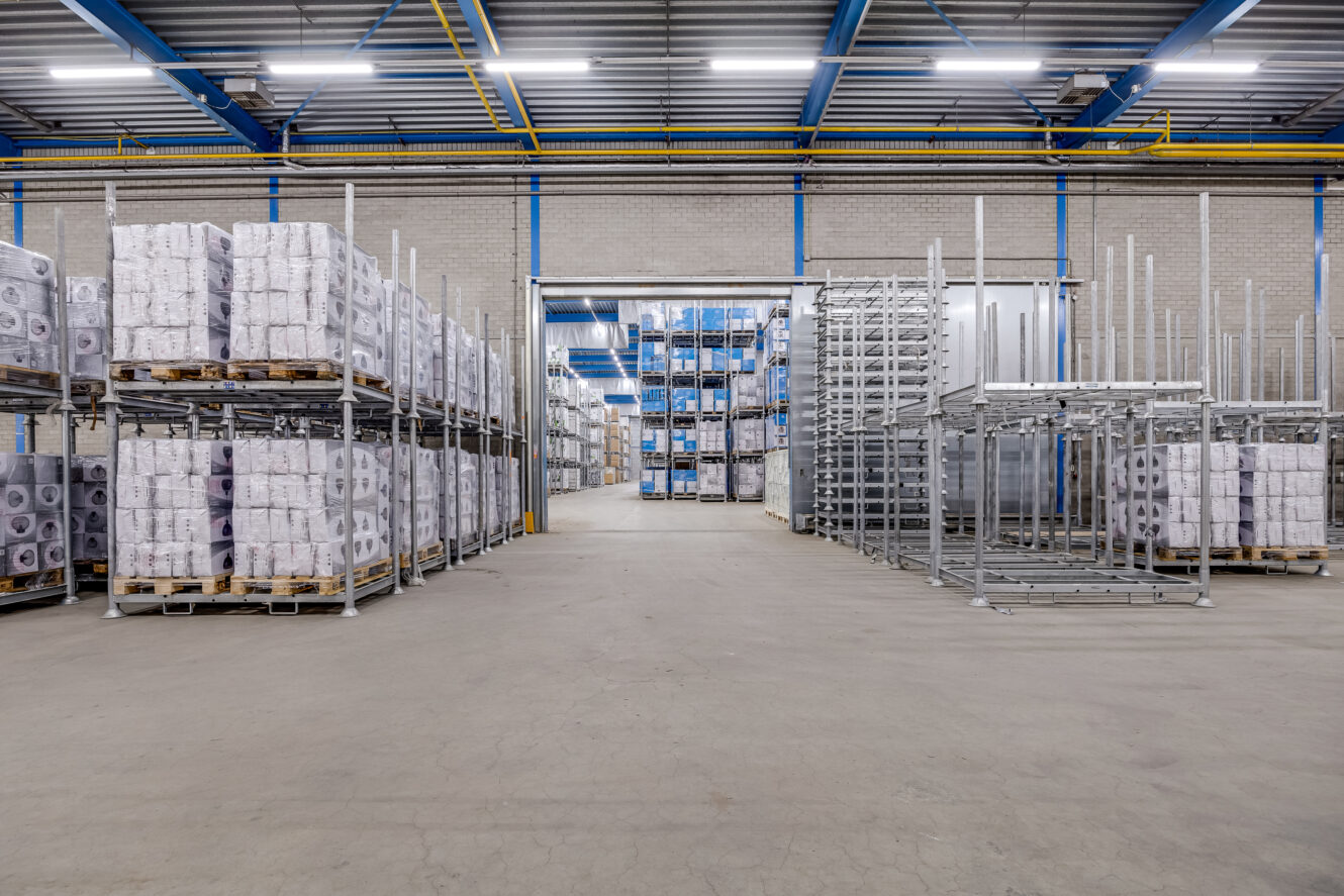 Groot magazijninterieur met gestapelde pallets met goederen aan de linkerkant, lege metalen rekken aan de rechterkant en een open deuropening die naar meer opslagruimte leidt.