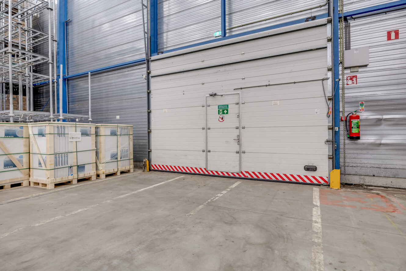 Grote industriële magazijndeur met bordje nooduitgang, omringd door metalen muren en pallets, met brandblusser en veiligheidsmarkeringen in de buurt.