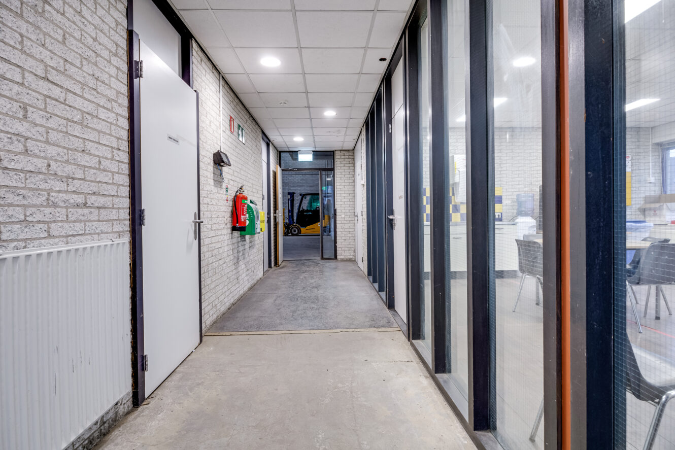 Een gang met bakstenen muren, een witte deur links, glazen ramen rechts en een brandblusser aan de muur. Een vorkheftruck is zichtbaar door de deuropening aan het einde.