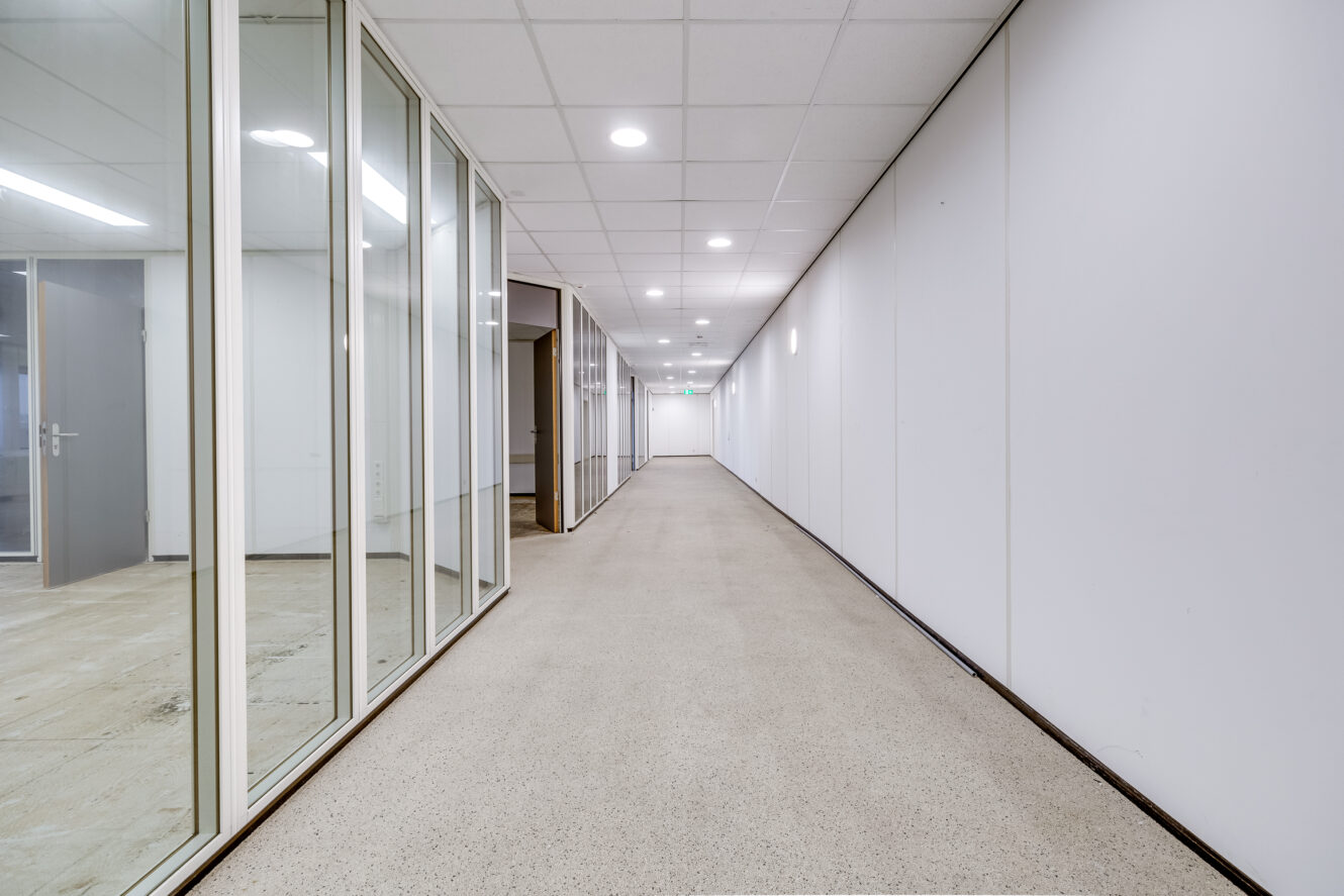 Een lange, lege gang met witte muren, een betegeld plafond en glazen kantoordeuren aan de linkerkant. De vloer is bedekt met lichtgekleurd linoleum. De gang is helder verlicht.