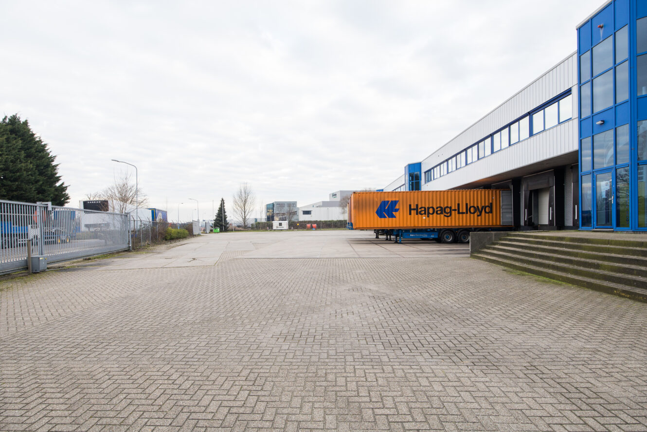 Een Hapag-Lloyd zeecontainer op een vrachtwagenaanhanger staat geparkeerd naast een modern industrieel gebouw met blauwomrande ramen en een leeg geplaveid terrein.