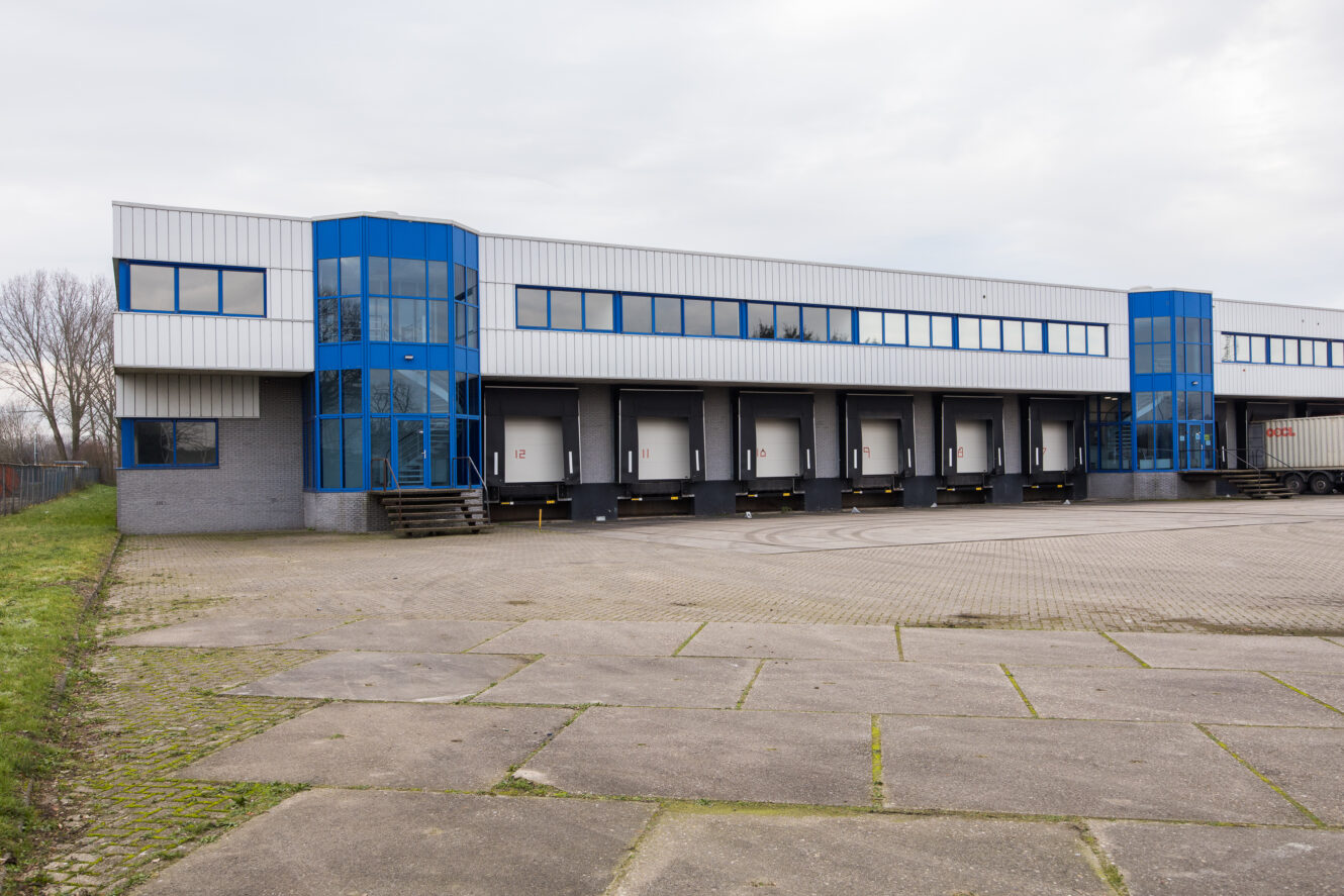 Een modern magazijngebouw met blauw omrande ramen en meerdere laadperrondeuren, gelegen naast een leeg geplaveid terrein onder een bewolkte hemel.