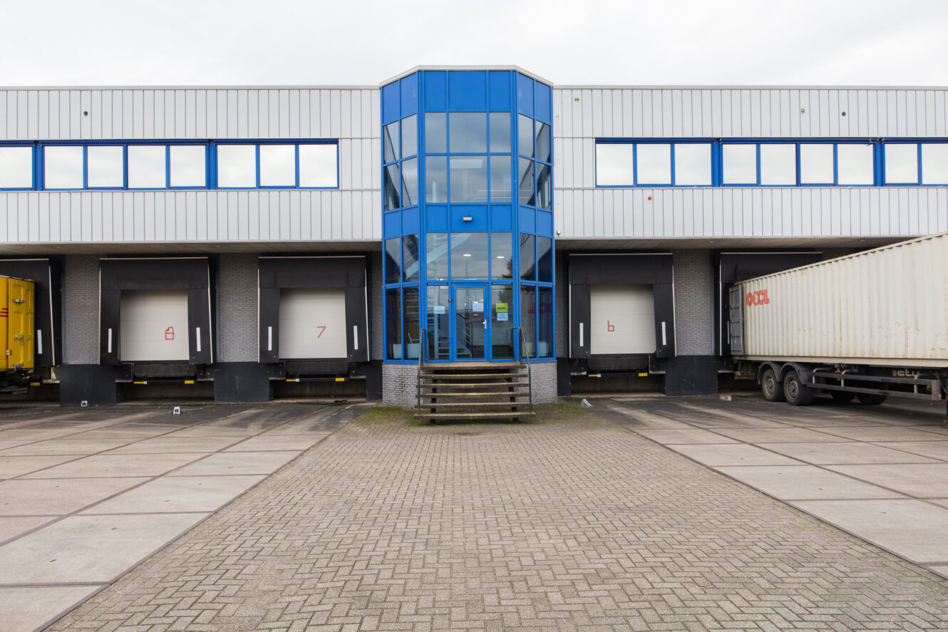 Een modern industrieel magazijn met een centrale ingang van blauw glas, vier laadkades en een grote witte vrachtwagen die rechts geparkeerd staat.
