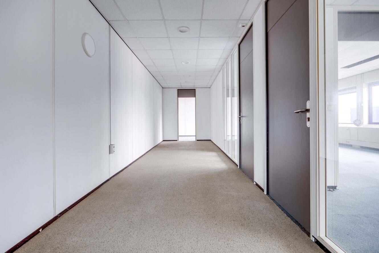 Lege gang met beige tapijt, witte muren en verschillende gesloten bruine en witte deuren; plafond met ingebouwde verlichting en een raam aan het einde van de gang.
