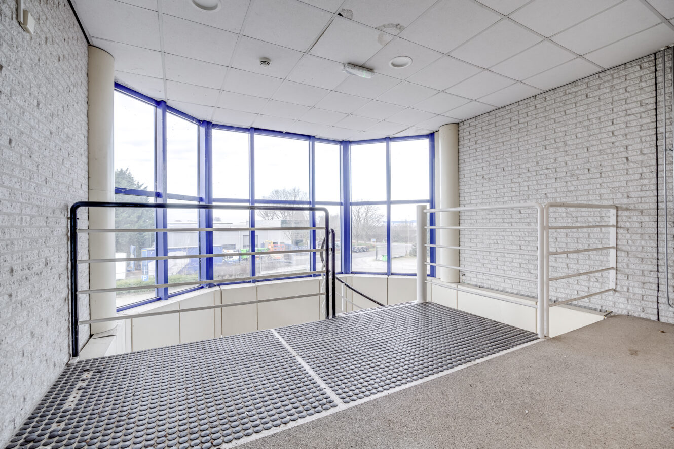 Overloop binnen met metalen roostervloeren en witte reling, naast grote ramen en bakstenen muren, met uitzicht op buiten.