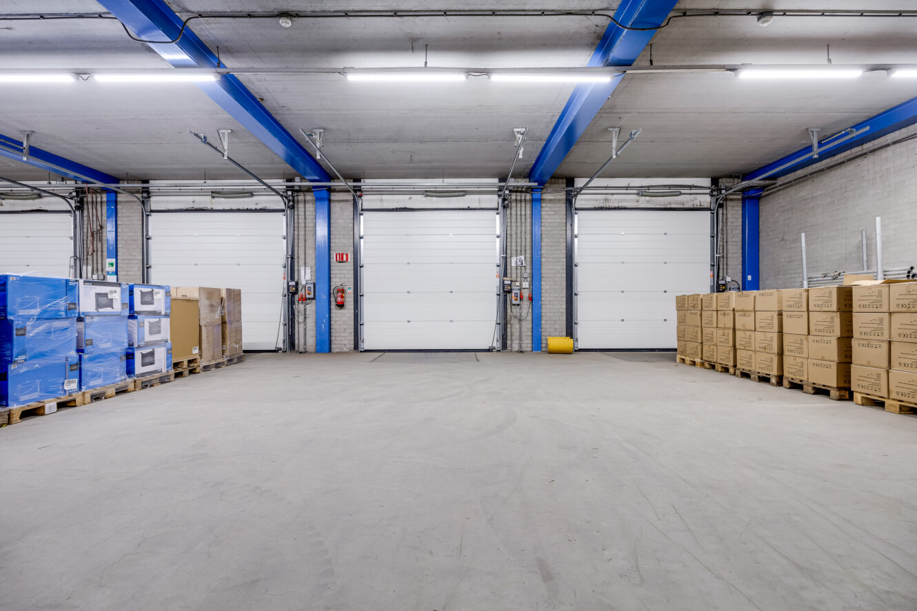 Magazijninterieur met gestapelde pallets met dozen aan beide zijden, betonnen vloer en drie grote gesloten laadperrondeuren aan de achterkant.