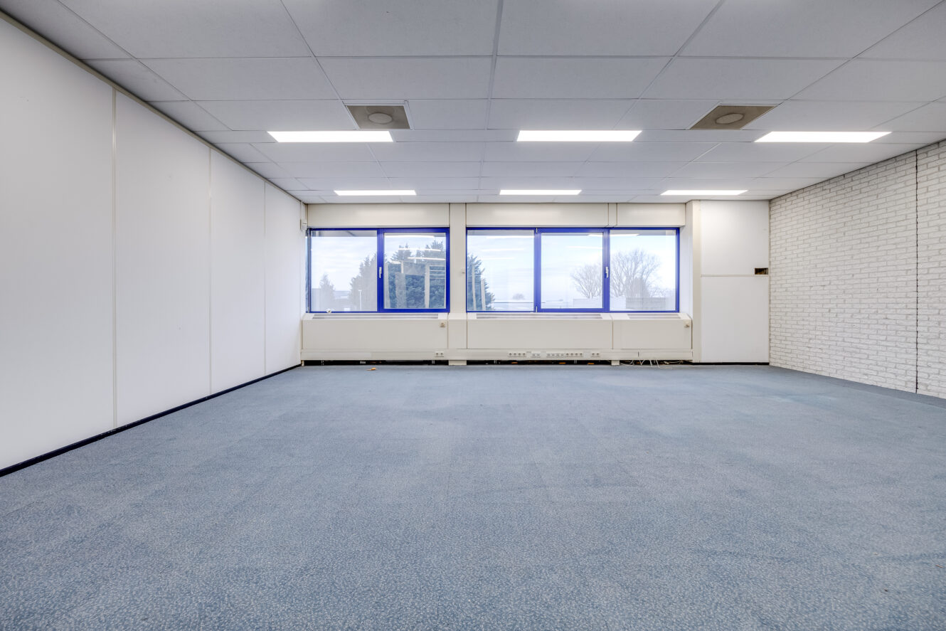 Lege kantoorkamer met blauw tapijt, witte muren, grote ramen en fluorescerende plafondverlichting.