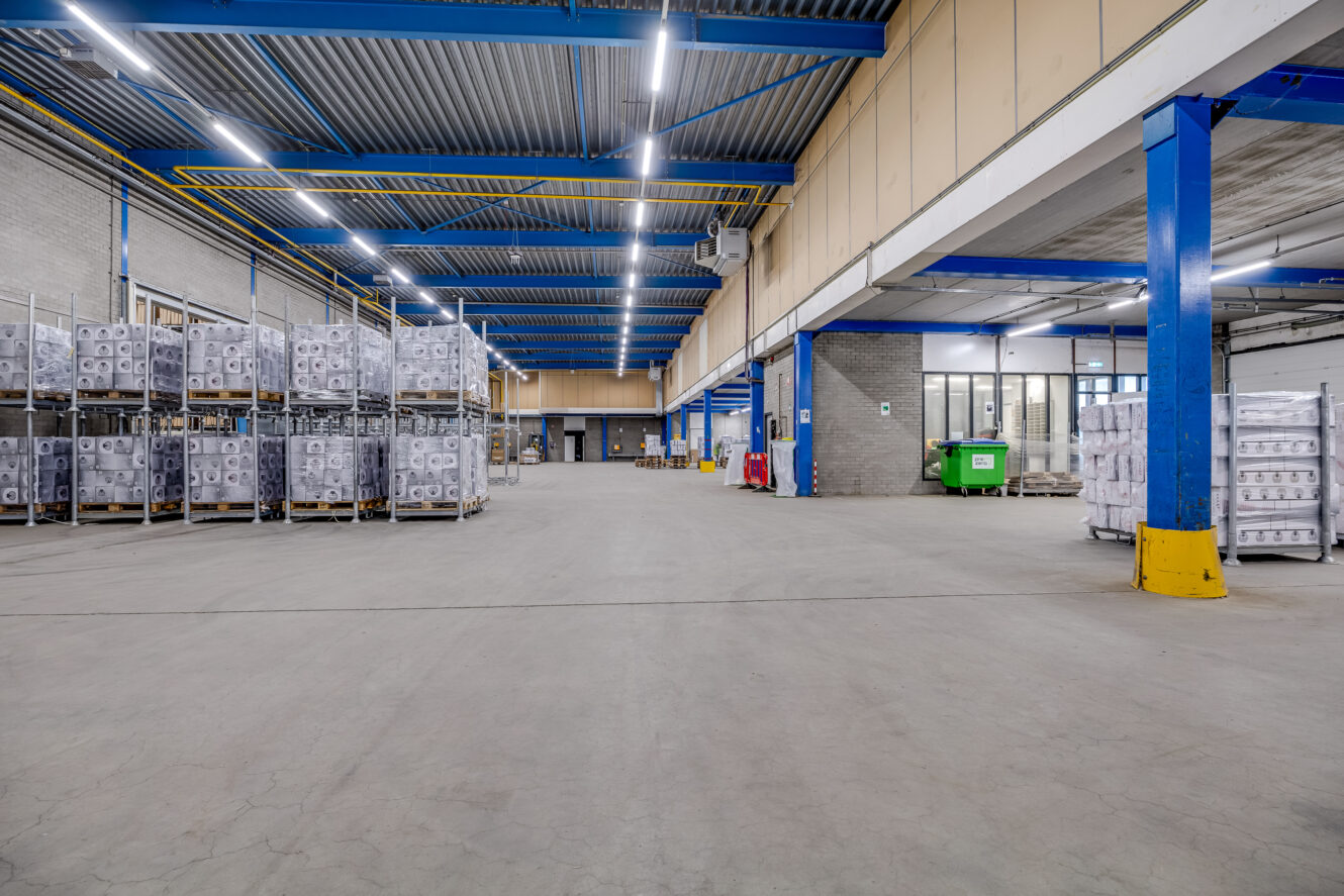 Groot magazijninterieur met hoge plafonds, blauwe stalen balken en stapels goederen op metalen planken langs de muren; een open ruimte in het midden.