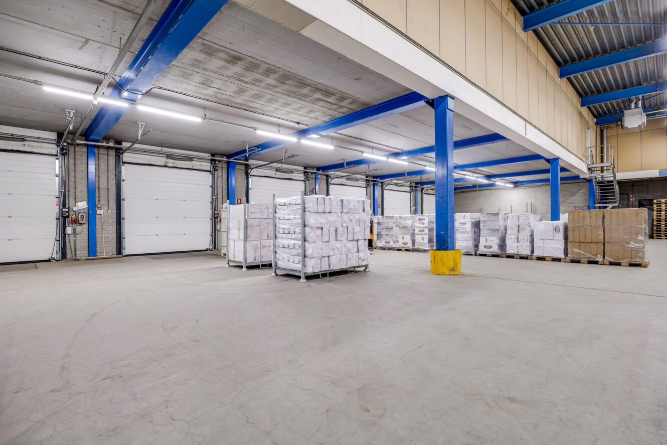 Ruim magazijninterieur met blauwe steunbalken, gestapelde pallets met goederen, grote roldeuren en heldere bovenverlichting.