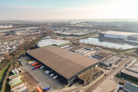 Luchtfoto van een groot industrieel magazijn omringd door geparkeerde vrachtwagens, laadkades, wegen en nabijgelegen commerciële gebouwen.
