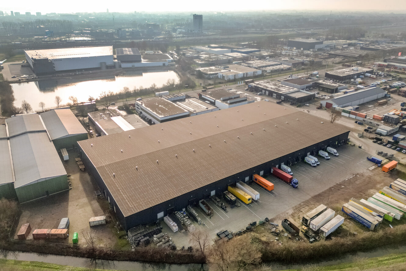Luchtfoto van een groot magazijn met meerdere vrachtwagens geparkeerd bij laadkades, omringd door industriële gebouwen en opslagcontainers.