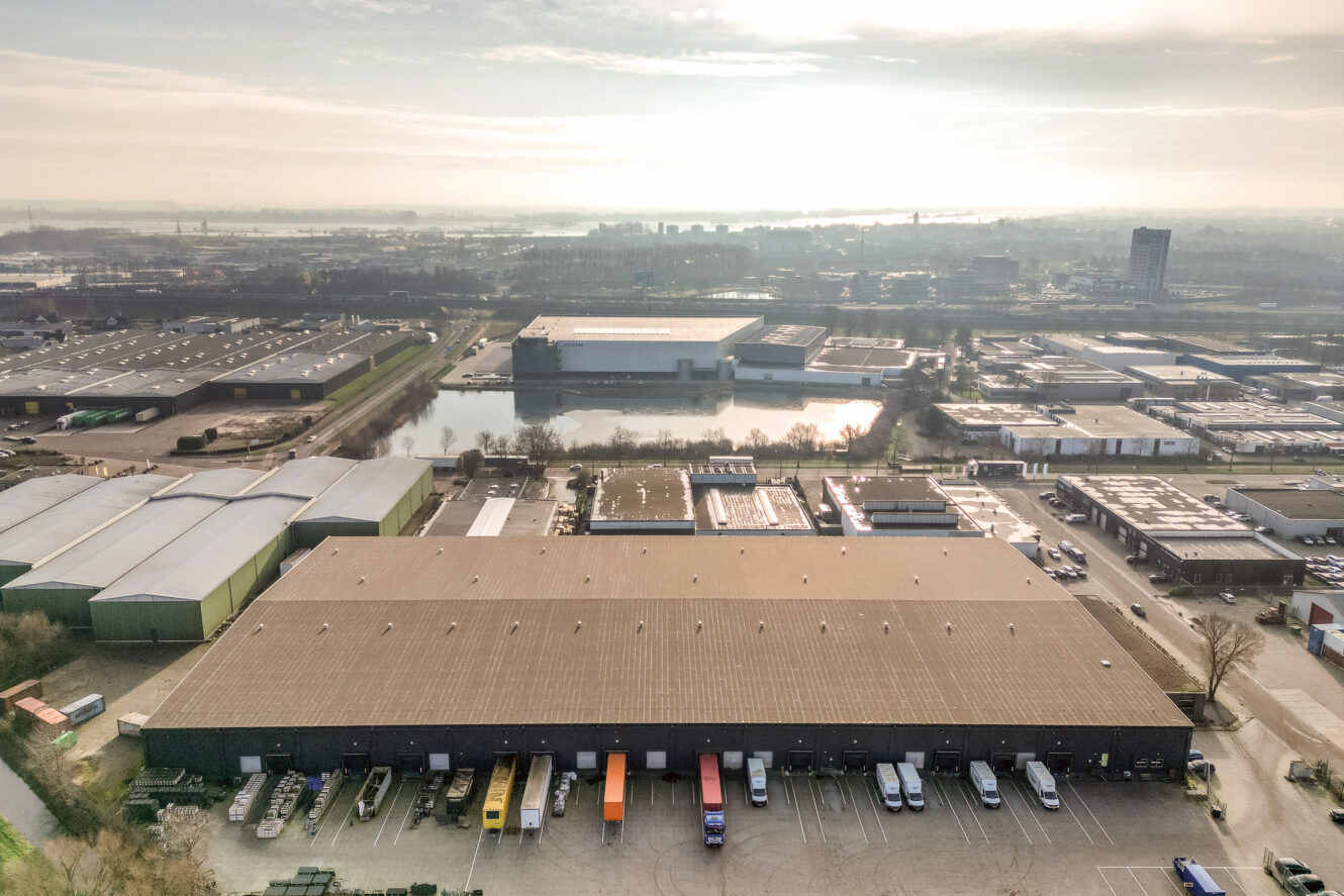 Luchtfoto van een industriegebied met magazijnen, geparkeerde vrachtwagens bij laadkades, verharde wegen en een vijver omringd door gebouwen onder een bewolkte hemel.