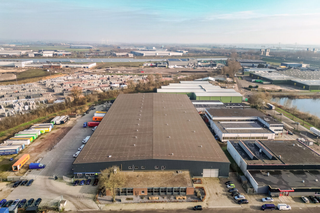 Luchtfoto van een groot magazijngebouw omringd door vrachtwagens, industriële eenheden en open land op een heldere dag.