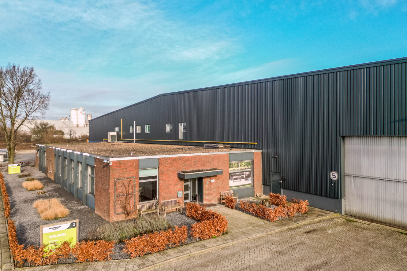 Een modern industrieel gebouw met een bakstenen kantoorgedeelte aan de voorkant en een groot zwart magazijn aan de achterkant, omringd door aangelegde struiken en een verharde oprit.