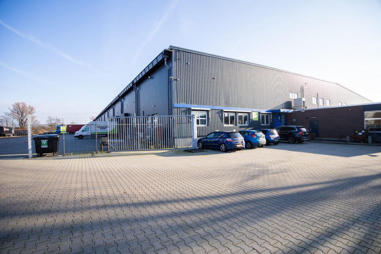 Een groot industrieel pakhuis met metalen gevelbekleding en een omheind parkeerterrein met verschillende auto's op een heldere, zonnige dag.