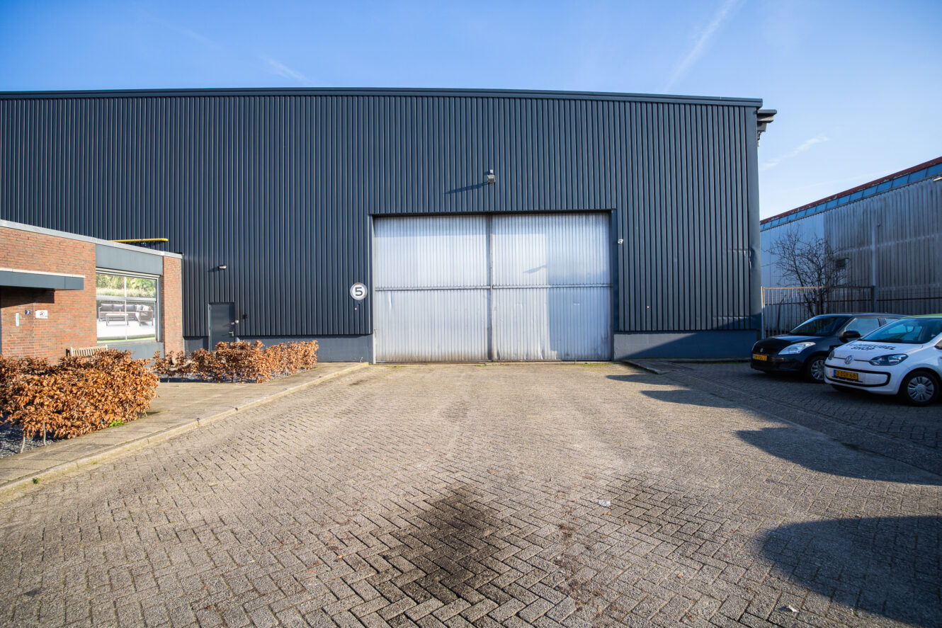 Een groot industrieel pakhuis met een gesloten metalen deur, aangrenzend bakstenen gebouw, geparkeerde auto's en een heldere lucht boven je hoofd.
