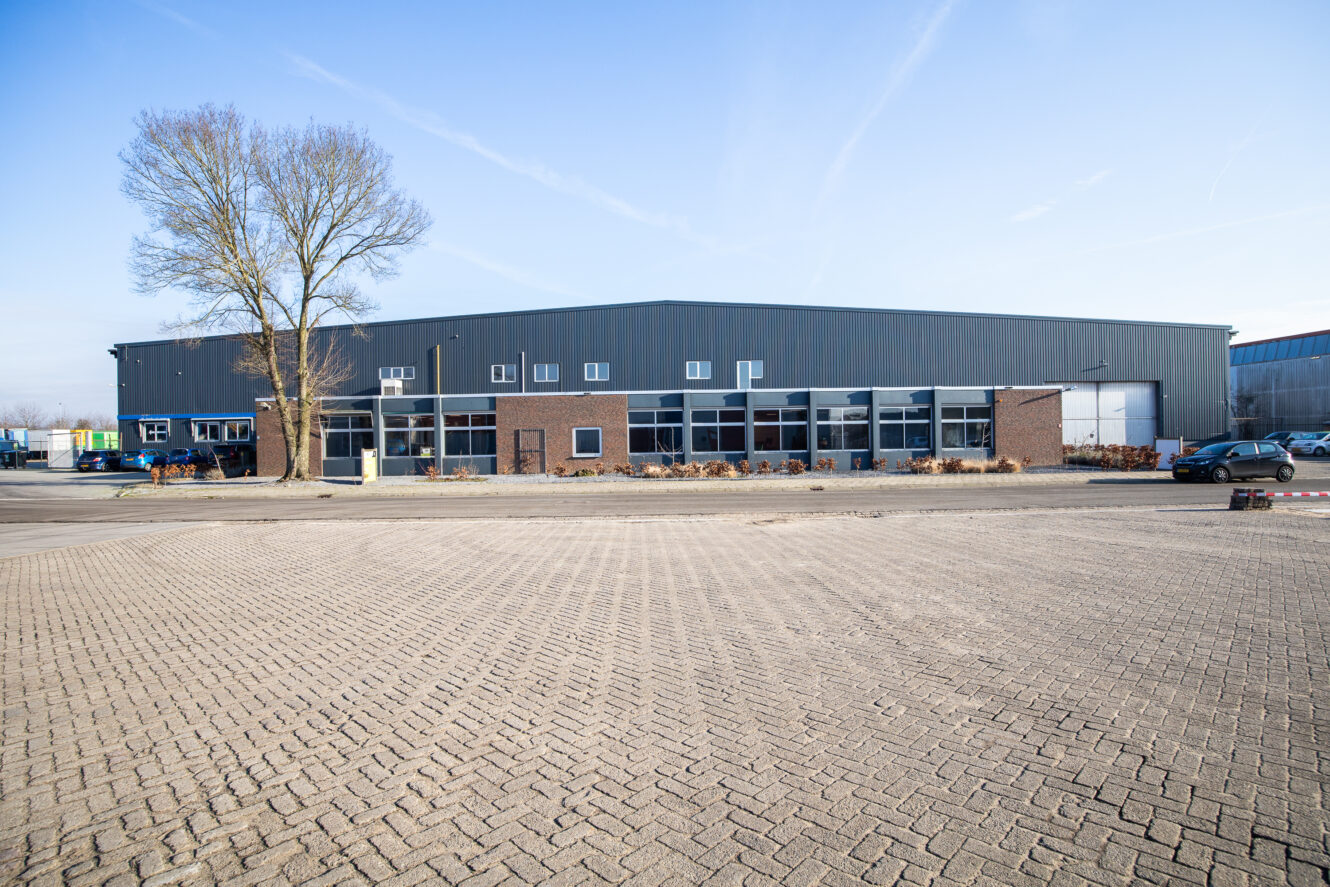 Een groot industrieel pakhuis met een mix van bakstenen en metalen muren, meerdere ramen en een geplaveide parkeerplaats aan de voorkant onder een strakblauwe hemel.