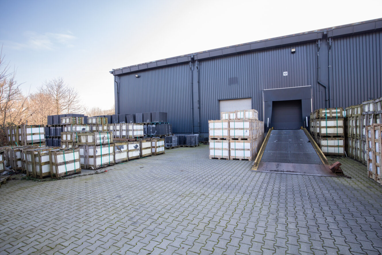 Industrieel laadgebied met gestapelde pallets en kratten, een metalen helling die leidt naar een grote gesloten deur en een geplaveid stenen oppervlak voor een grijs magazijngebouw.