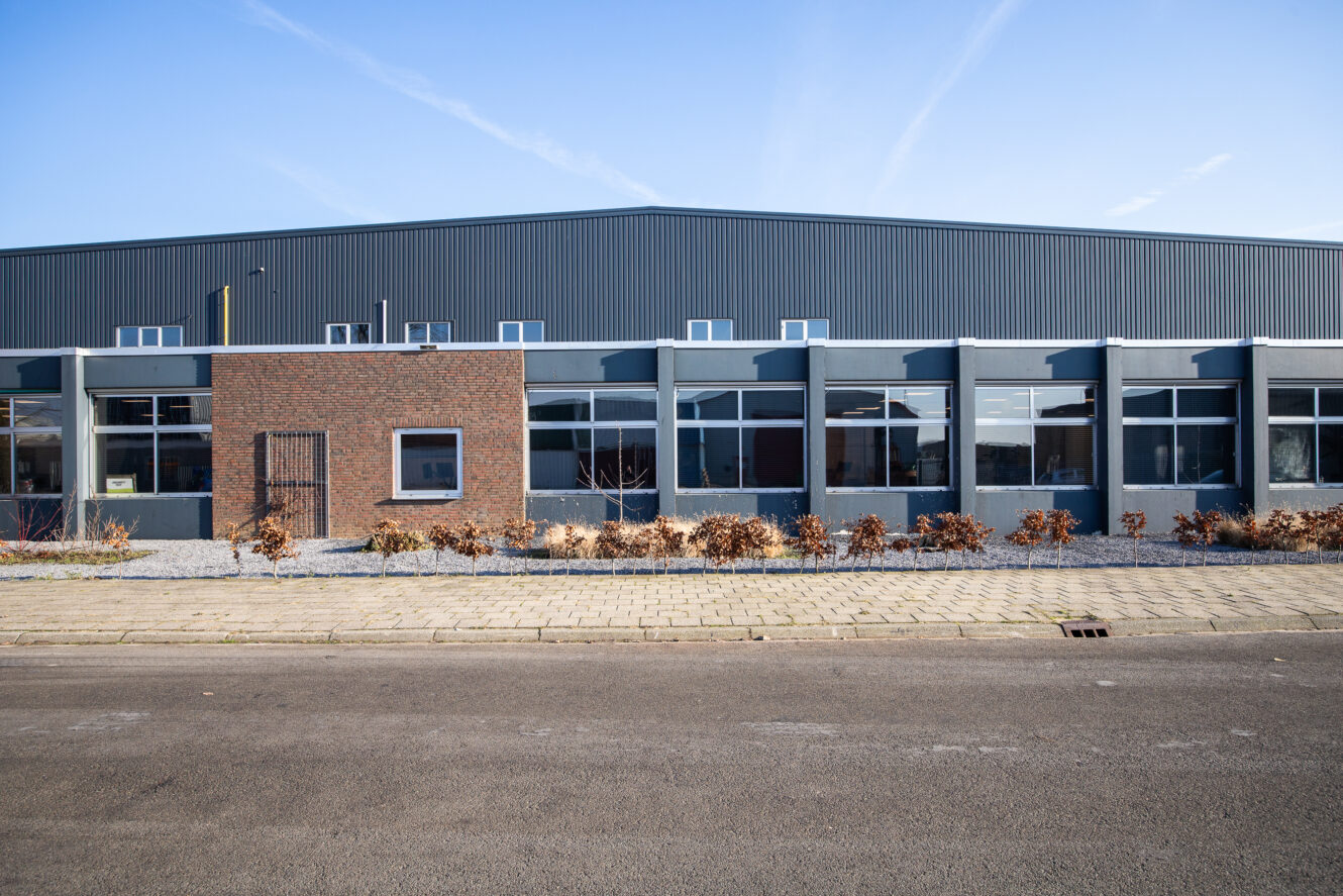 Een modern industrieel gebouw met grote ramen, een bakstenen en metalen gevel, lage struiken en een geplaveid trottoir onder een strakblauwe hemel.