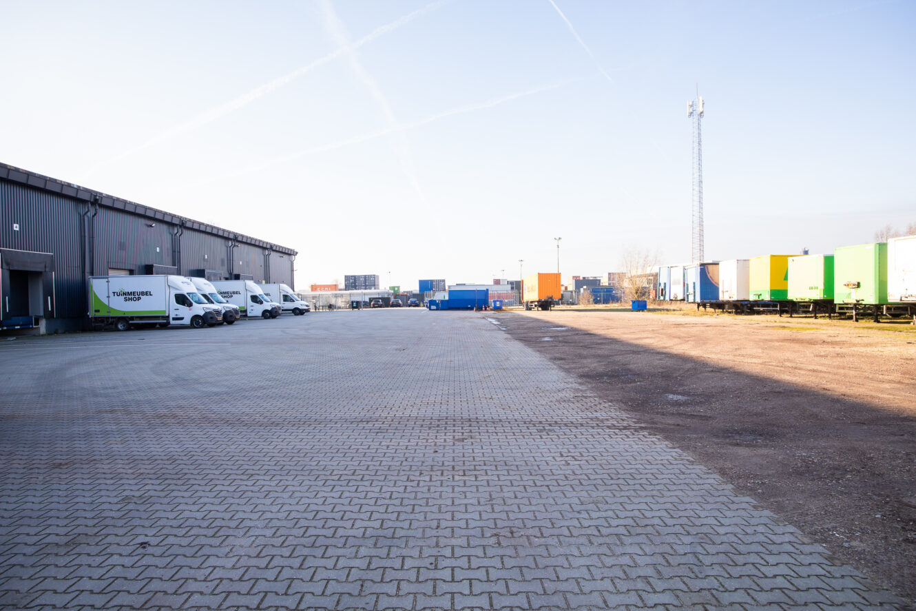 Een geasfalteerd terrein voor een magazijn met links verschillende bestelwagens geparkeerd en rechts verschillende gekleurde zeecontainers en opleggers.