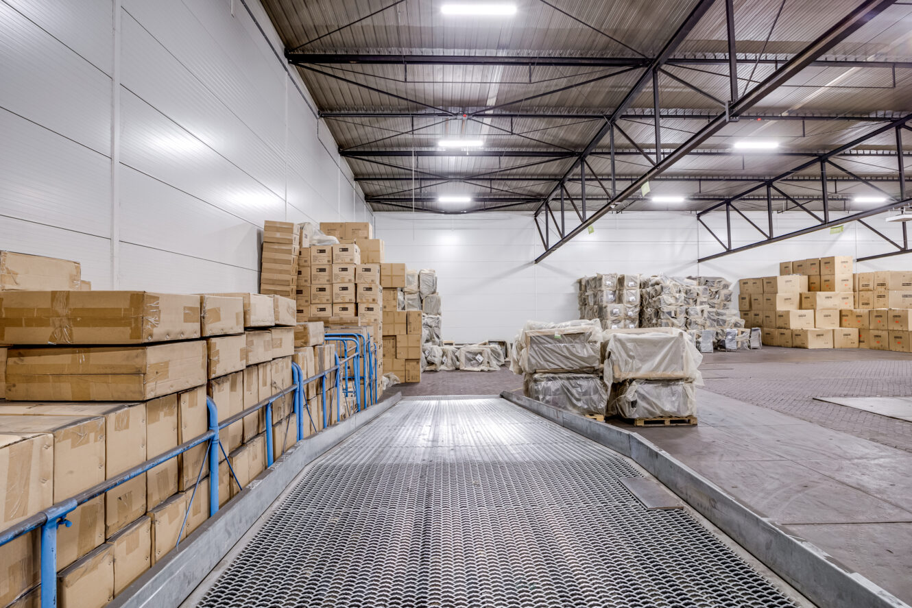 Interieur van een magazijn met stapels kartonnen dozen en pallets, metalen hellingbaan op de voorgrond en felle lampen aan het plafond.