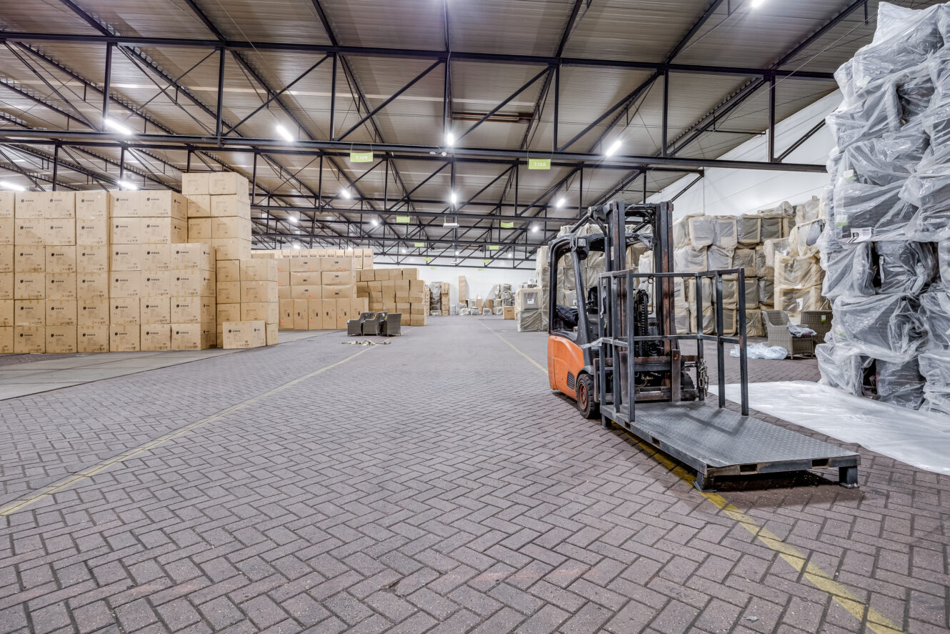 Een vorkheftruck staat geparkeerd in een groot magazijn met stapels kartonnen dozen en ingepakte pallets langs de muren.