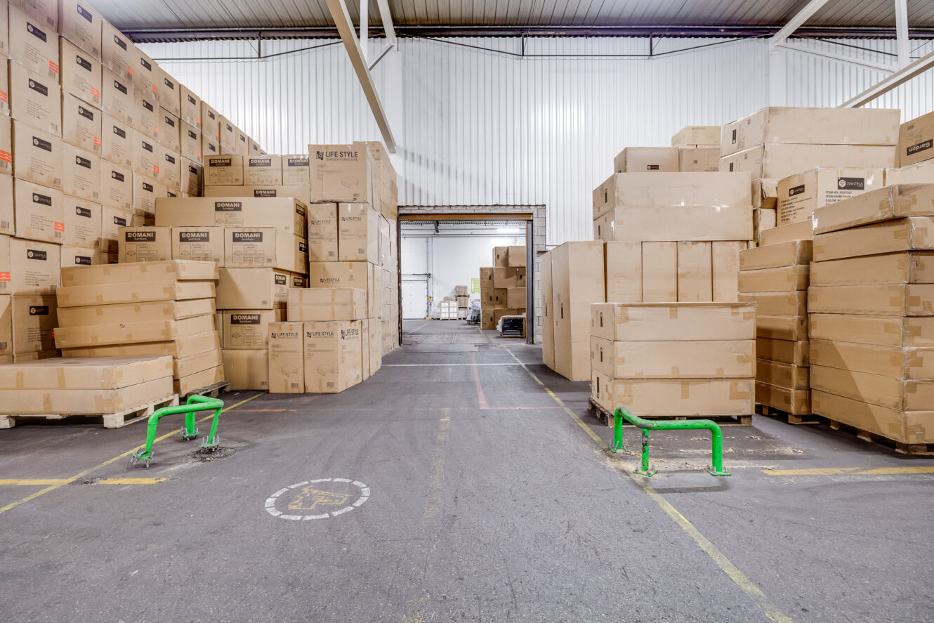 Stapels grote kartonnen dozen staan op pallets in een ruim magazijn met hoge plafonds en betonnen vloeren.