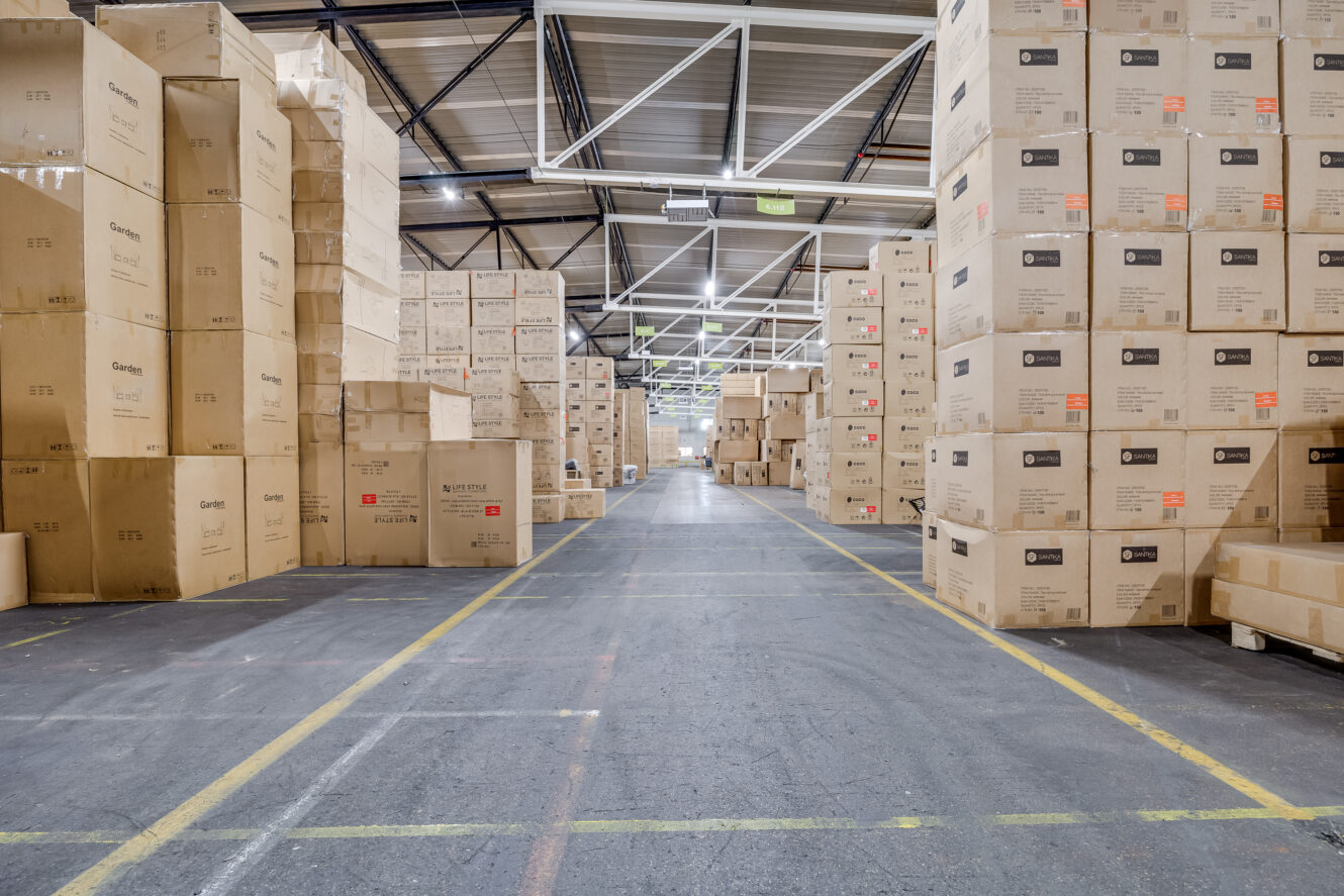 Groot magazijninterieur met hoge stapels kartonnen dozen in rijen langs een breed middenpad, onder heldere plafondverlichting.