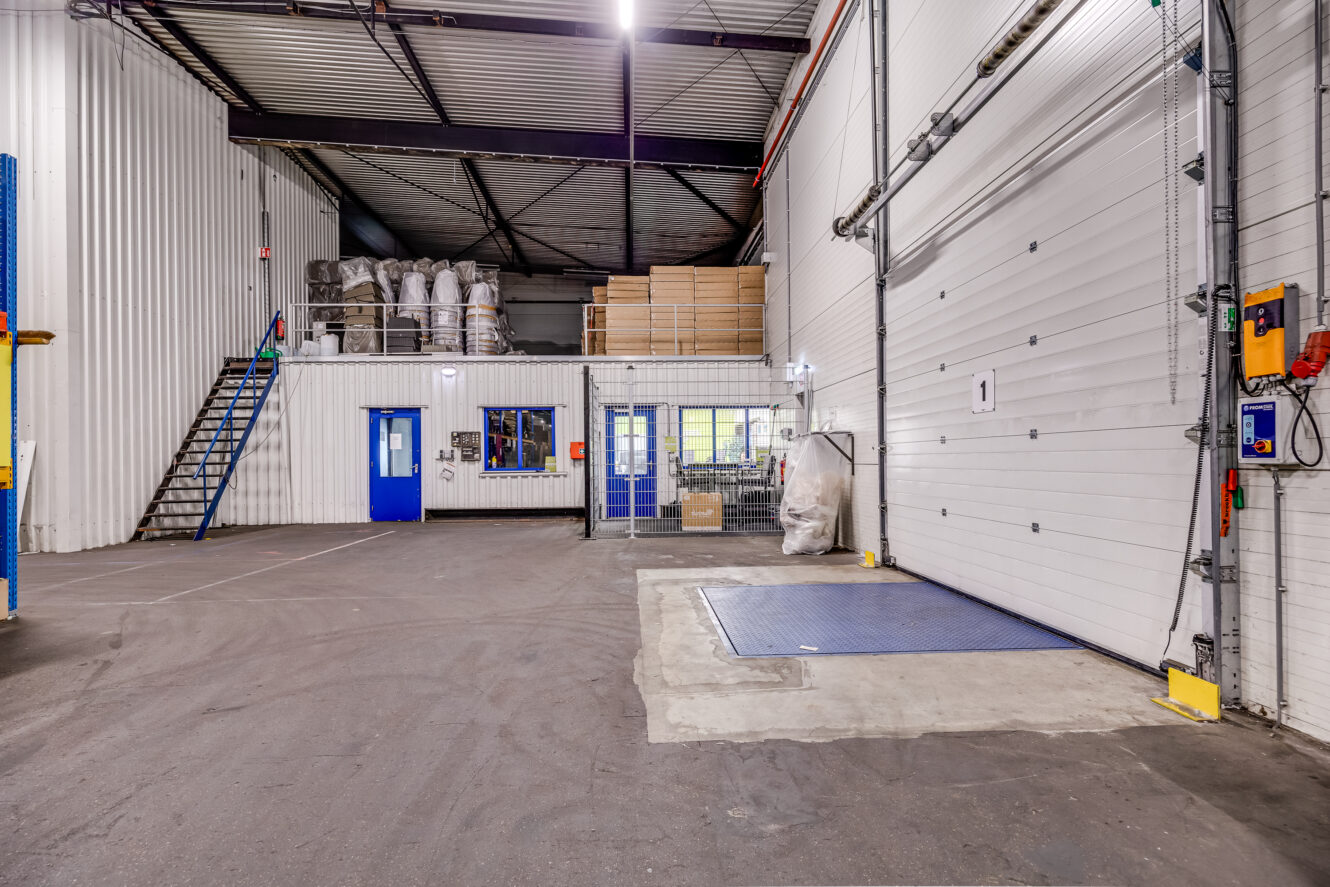Interieur van een magazijn met een grote laaddeur, een blauwe platformweegschaal, gestapelde pallets, trappen naar een zolder en kantoorruimtes met blauwe deuren en ramen.