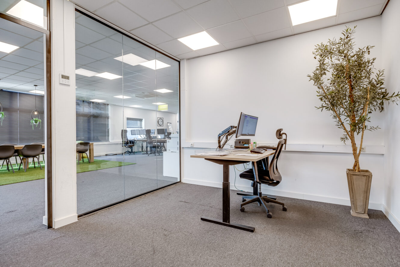 Modern kantoor met een enkel bureau, stoel, computer en plant in pot naast een glazen wand met uitzicht op een vergaderruimte met stoelen en tafels.