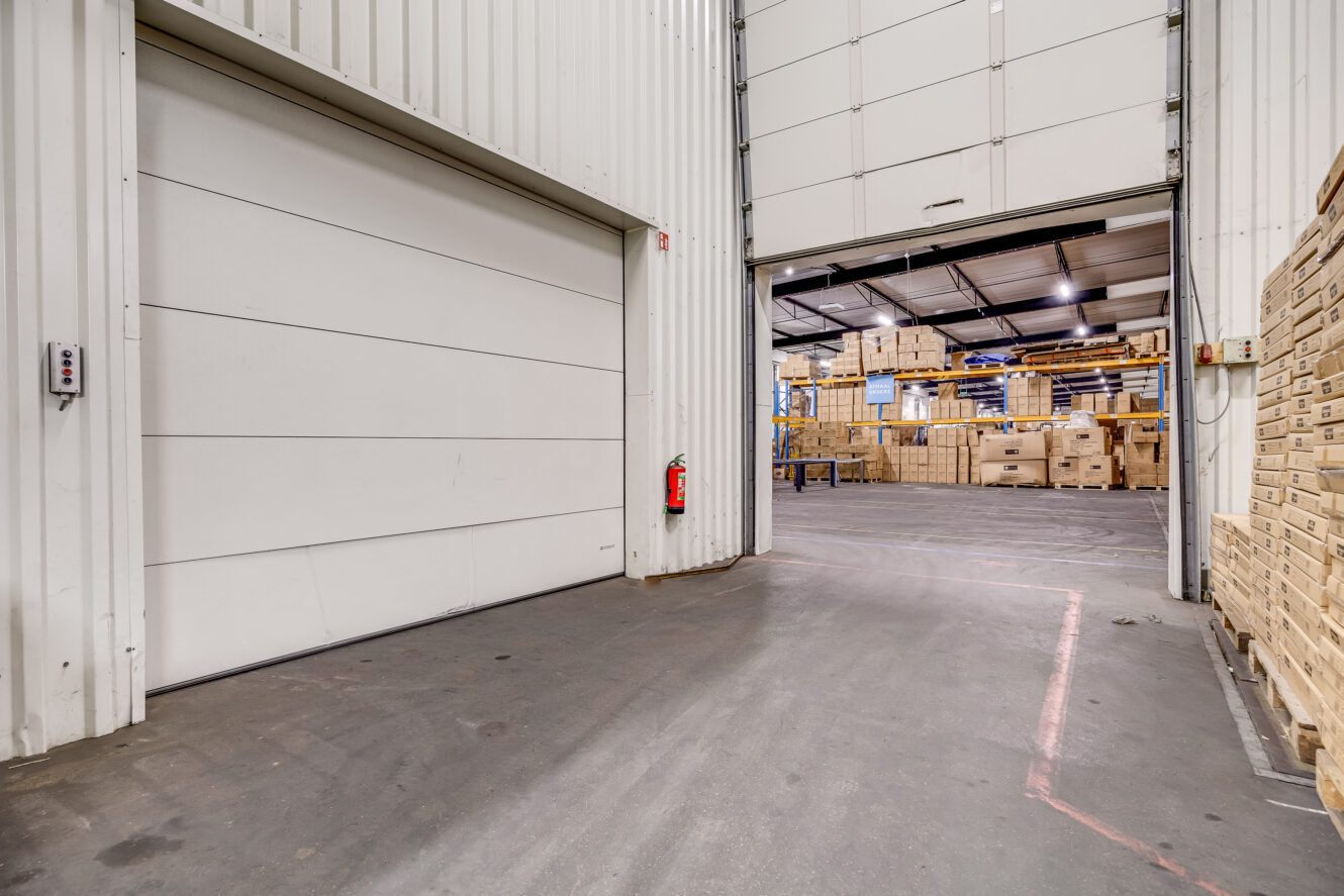 Interieur van een magazijn met gestapelde houten kratten, een gedeeltelijk openstaande grote deur en een brandblusser aan de muur.