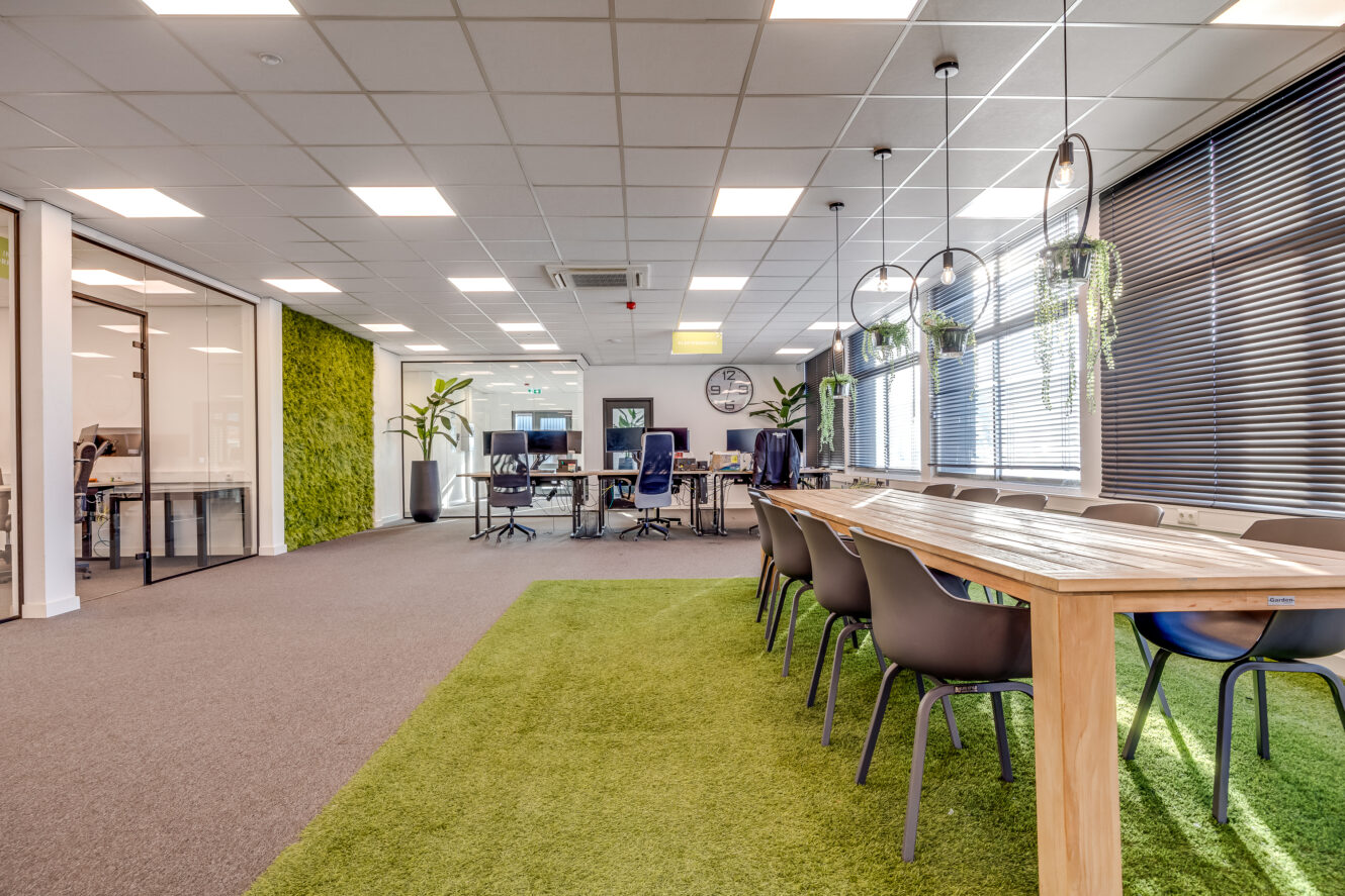 Moderne kantoorruimte met een lange houten tafel, zwarte stoelen, vloerbedekking en groene graszoden, planten en vergaderruimten met glazen wanden.