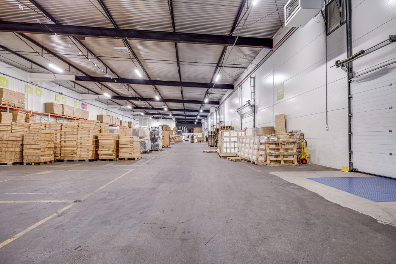 Groot magazijn met hoge plafonds, pallets met kisten, gestapelde houten kratten en planken langs de muren. Breed middenpad voor toegang voor voertuigen.