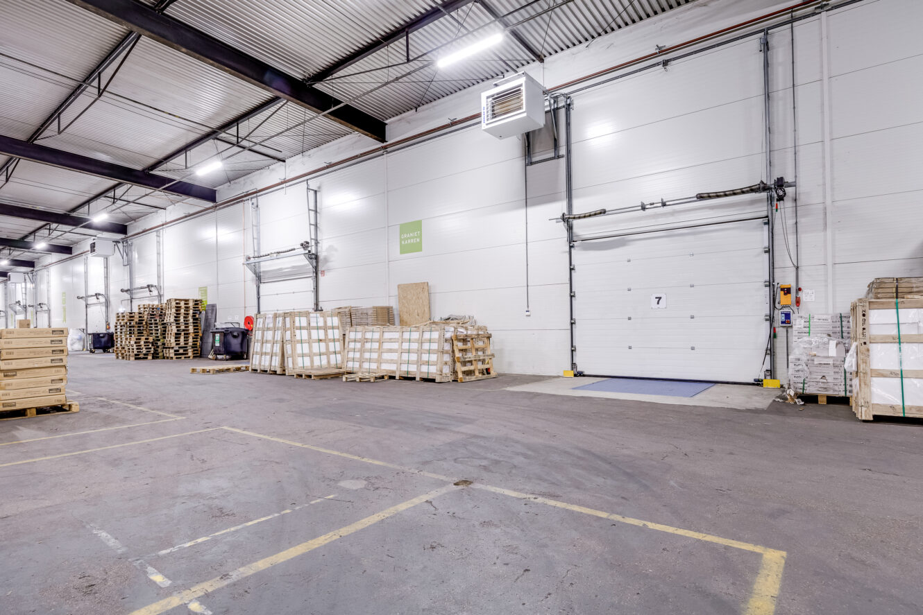 Interieur van een magazijn met gestapelde pallets, dozen en een grote witte laaddockdeur met het nummer 7 erop. De vloer is gemarkeerd met gele lijnen.