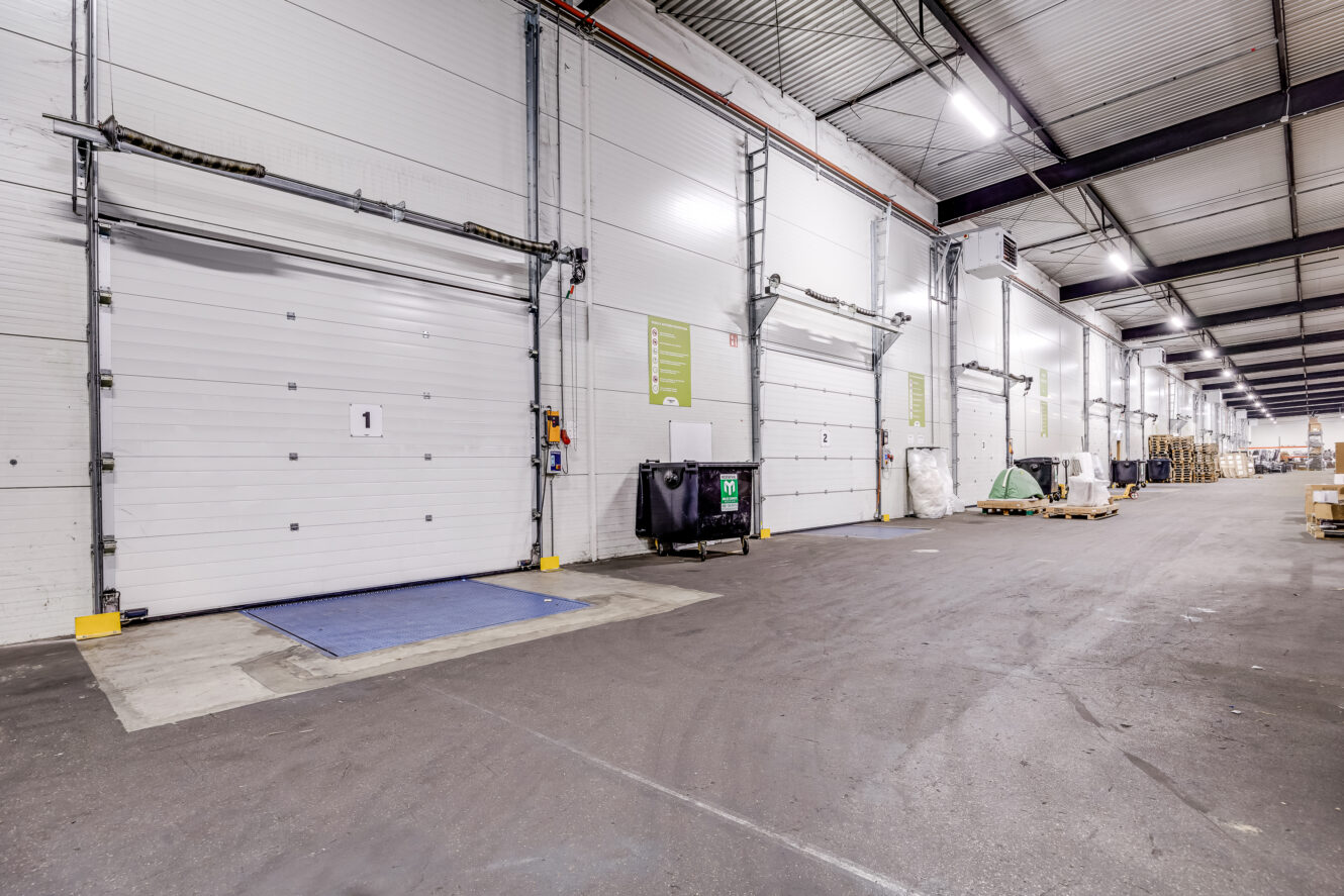 Binnenaanzicht van een magazijn met meerdere laaddockdeuren, een container, pallets en hoge plafonds met zichtbare verlichting.