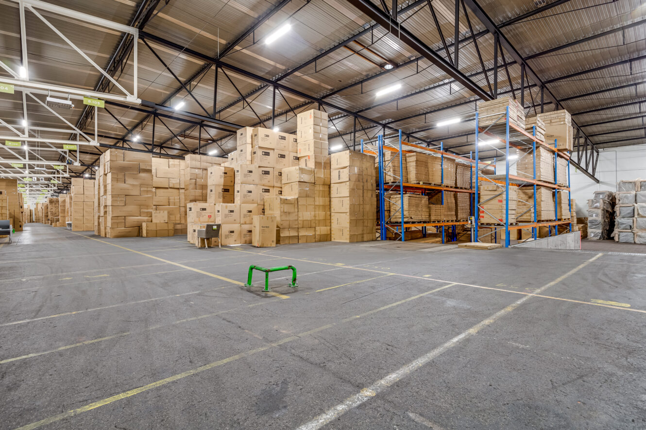 Groot magazijninterieur met stapels kartonnen dozen op pallets, lege rekken en overheadverlichting.