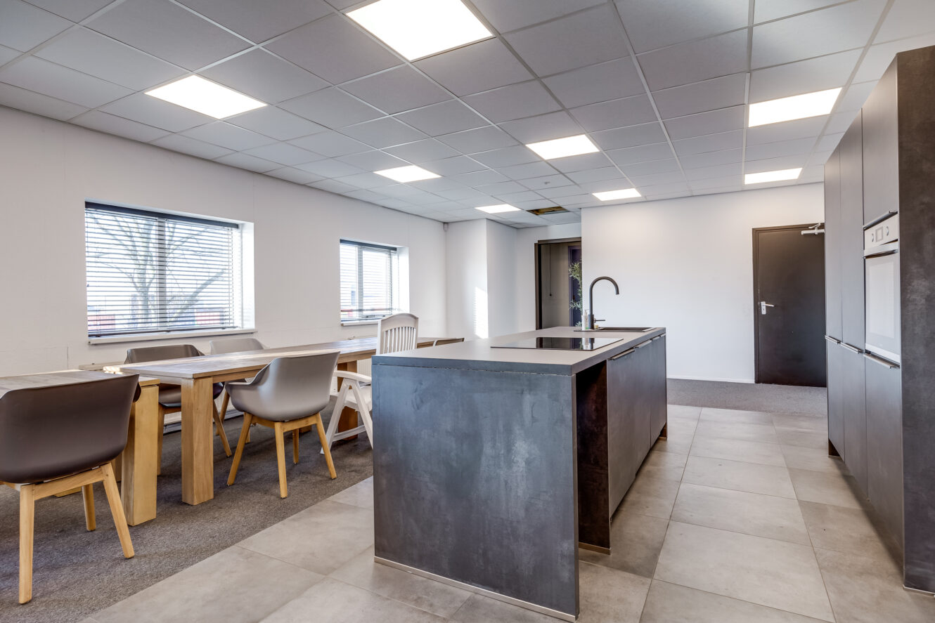 Moderne keuken met een centraal eiland, inbouwapparatuur, eettafel met gemengde stoelen, grote ramen en een tegelvloer onder een rasterplafond met tl-verlichting.